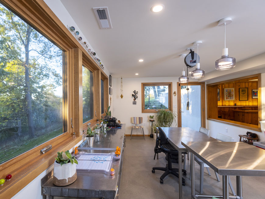 Simple art studio features medal work tables, views of property through a wall of windows, and a view into rec room.