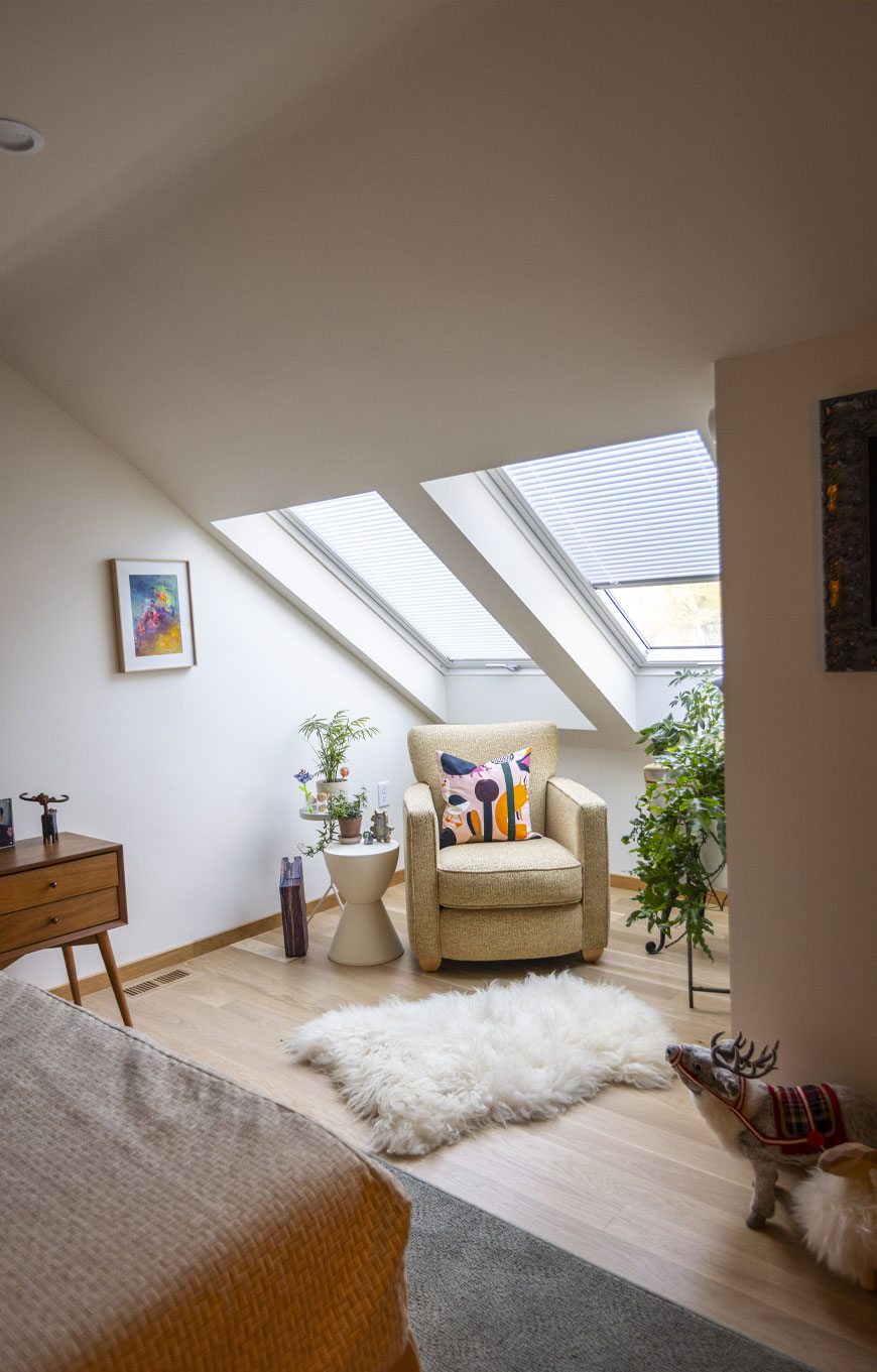 Sloped ceiling features skylights over a small reading nook to side of bedroom. A chair with brightly colored pillow and fluffy white rug sit under the windows with plants surrounding it.