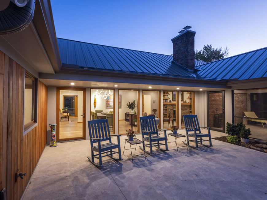 Night time shot of rear terrace of newly remodeled home in Kentucky by Kepes Architecture. Interior is lit from within, featuring white walls with ample art. Three rocking chairs sit at finished concrete terrace.