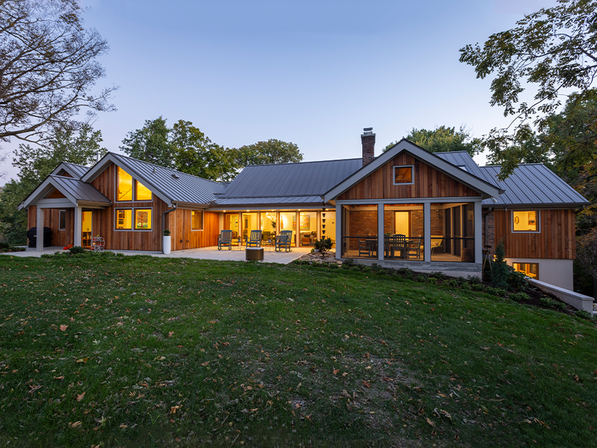 Back view of home remodeled by Kepes Architecture. Home is lit from within and emits a soft glow from windows. House has wooden siding and mid-century modern design.
